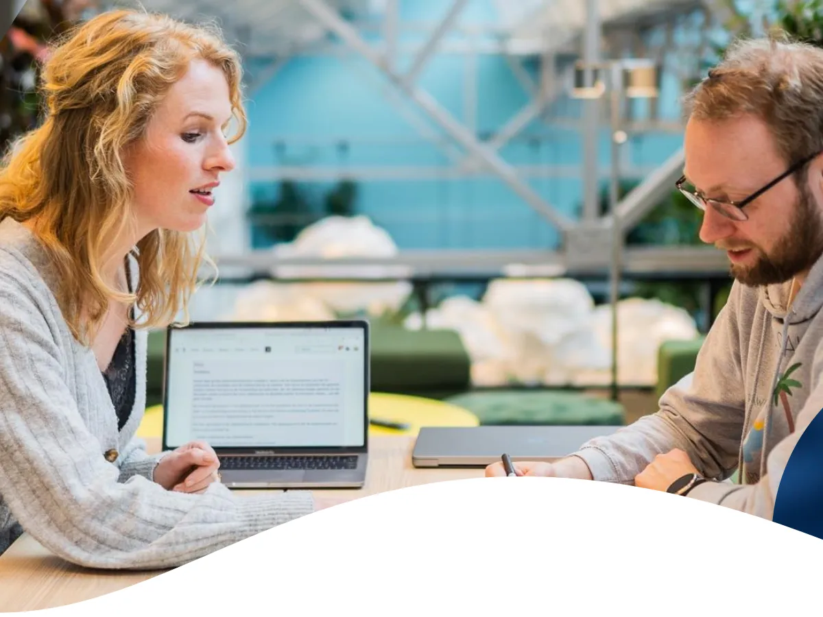2 mensen aan het werk aan tafel met een laptop op de achtergrond